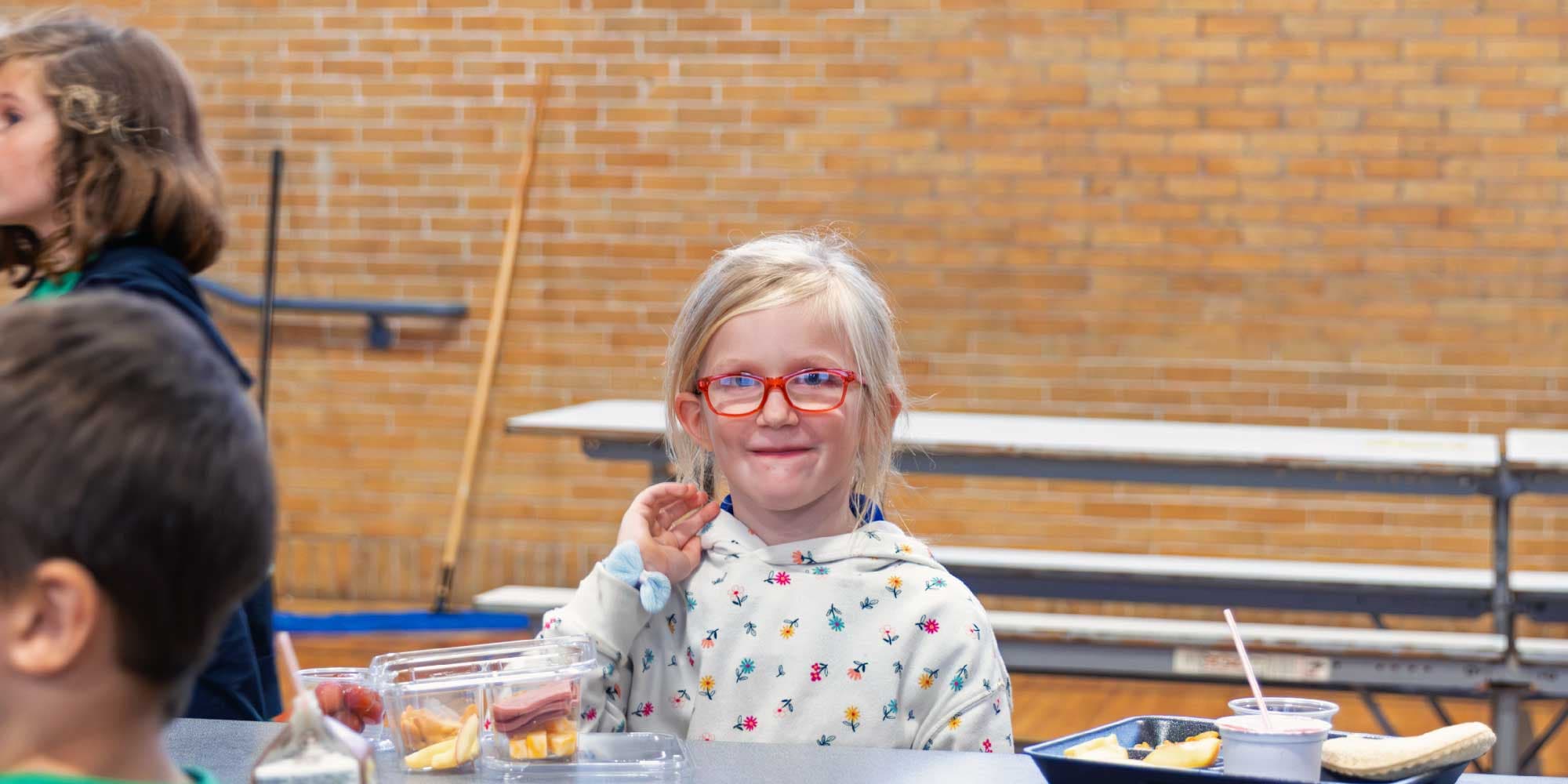 Student in red glasses smiling at a cafeteria lunch table.