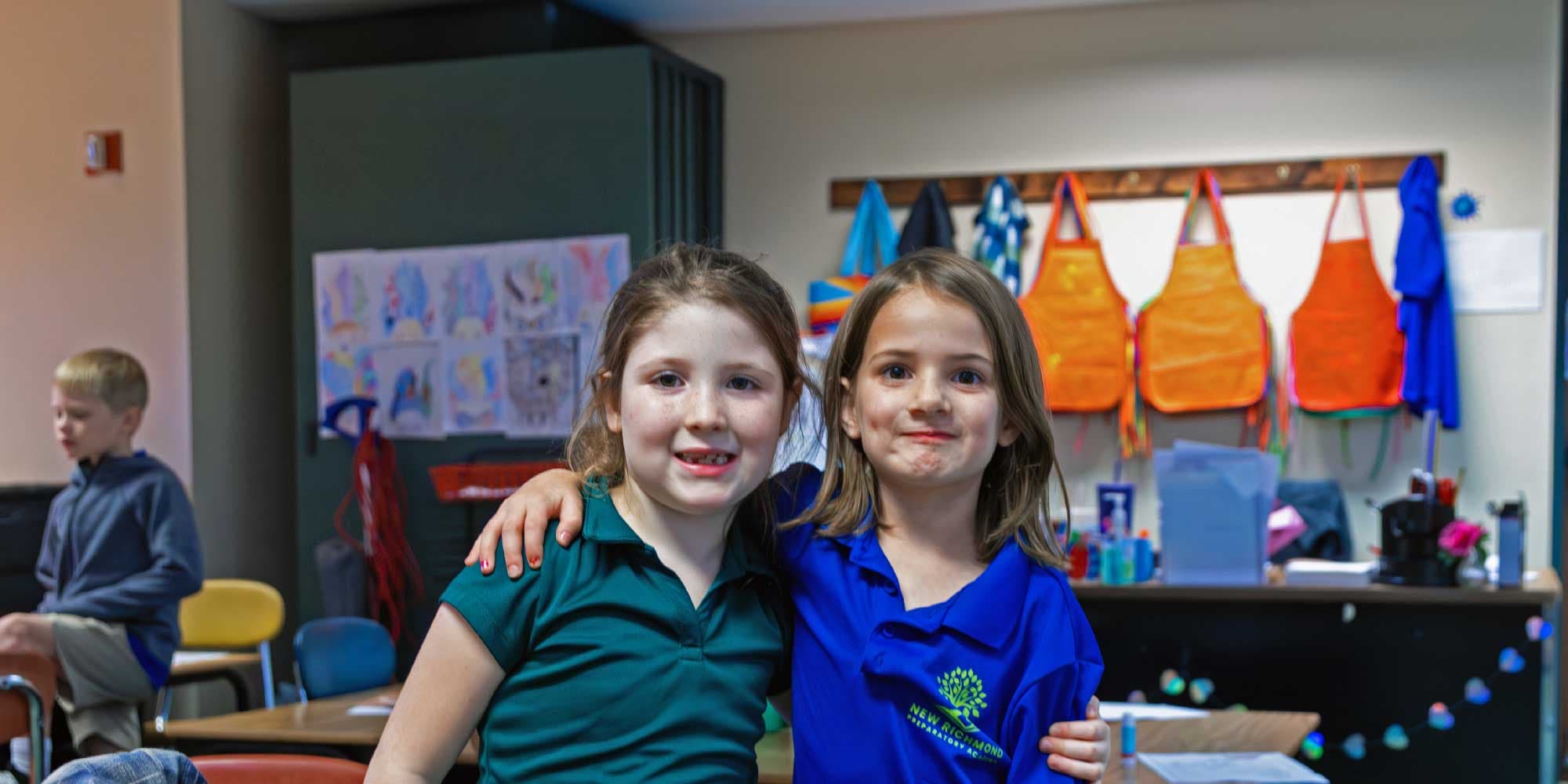Two smiling students pose arm in arm in a classroom.