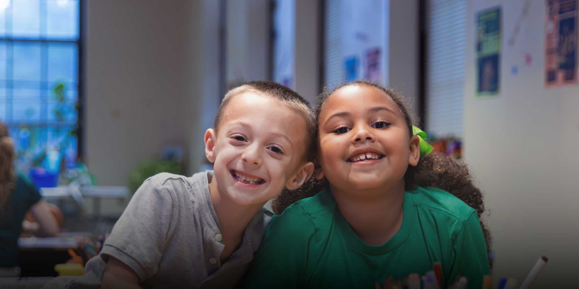 Two smiling students leaning together in a classroom.