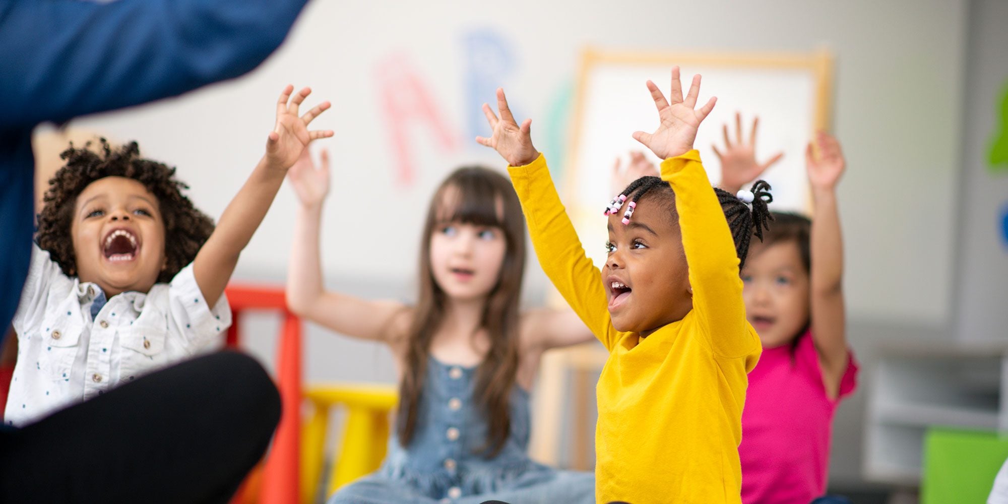 Four excited children raising hands in a classroom.