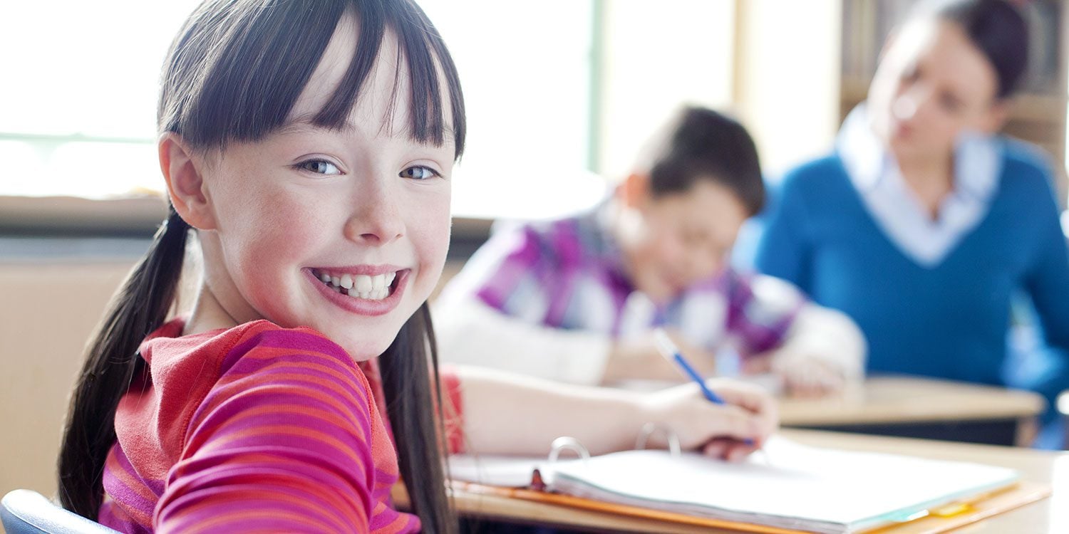 Smiling child in classroom with other students and an adult.